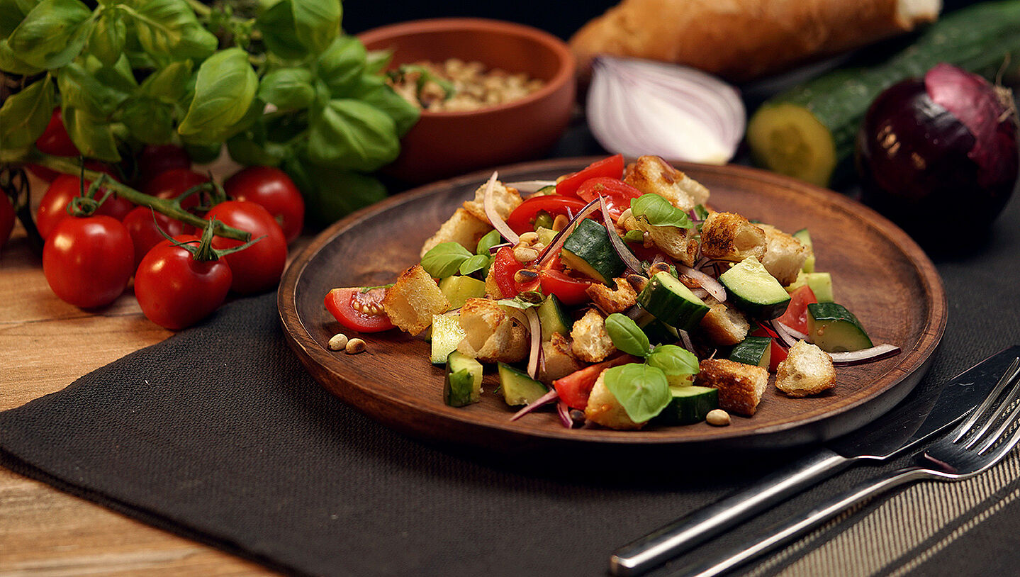 Auf einem Holzteller ist der italienischer Brotsalat namens Panzanella mit Gurken, Tomaten, Zwiebeln angerichtet