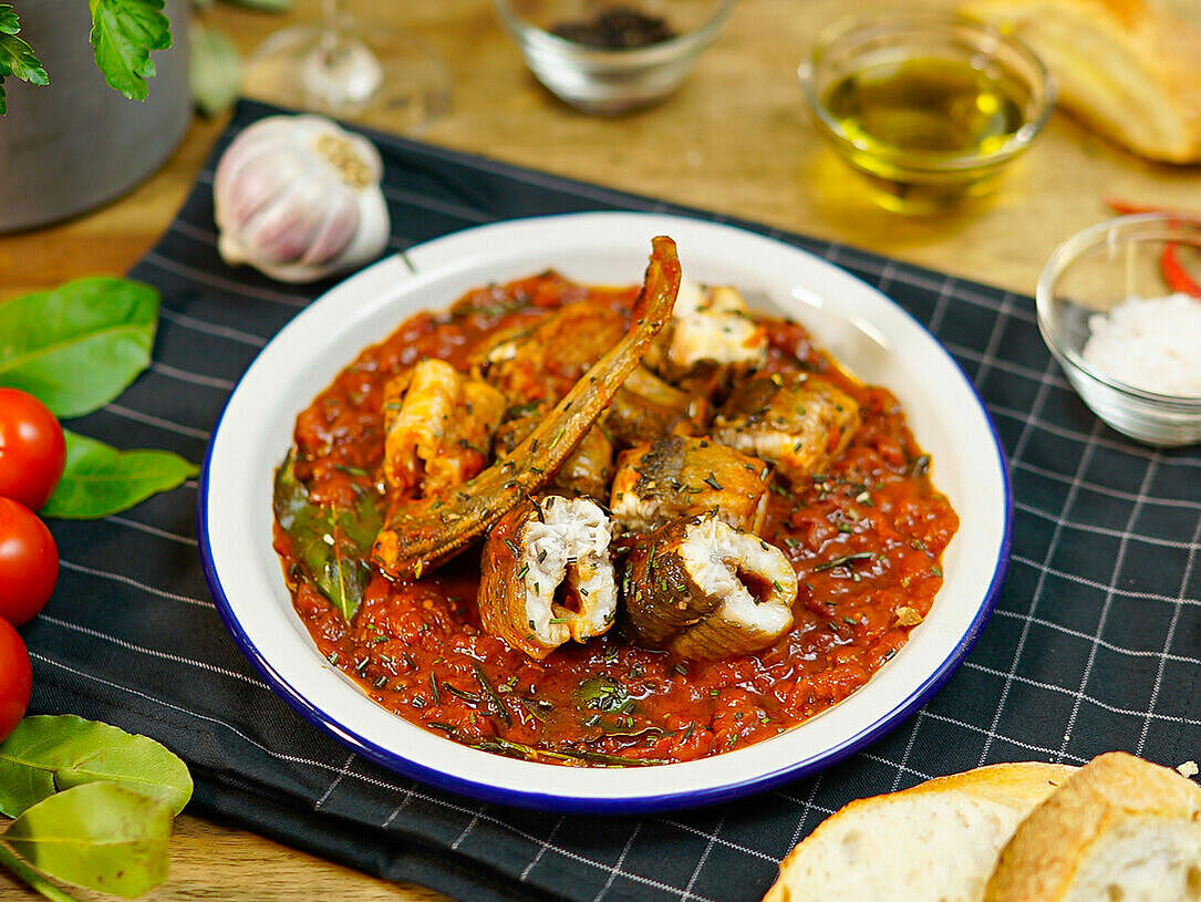 Auf einem weißen Teller mit blauen Rand ist ein traditionelles italienisches Aal-Rezept zu sehen. Der Aal liegt in Tomatensoße. Verschiedene frische Zutaten wie Tomaten, Basilikum sind als Deko zu sehen.