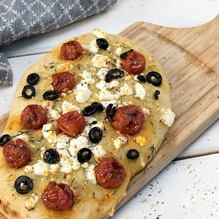 Auf einem Holzbrett liegt eine Focaccia mit Tomaten, Oliven und Schafskäse