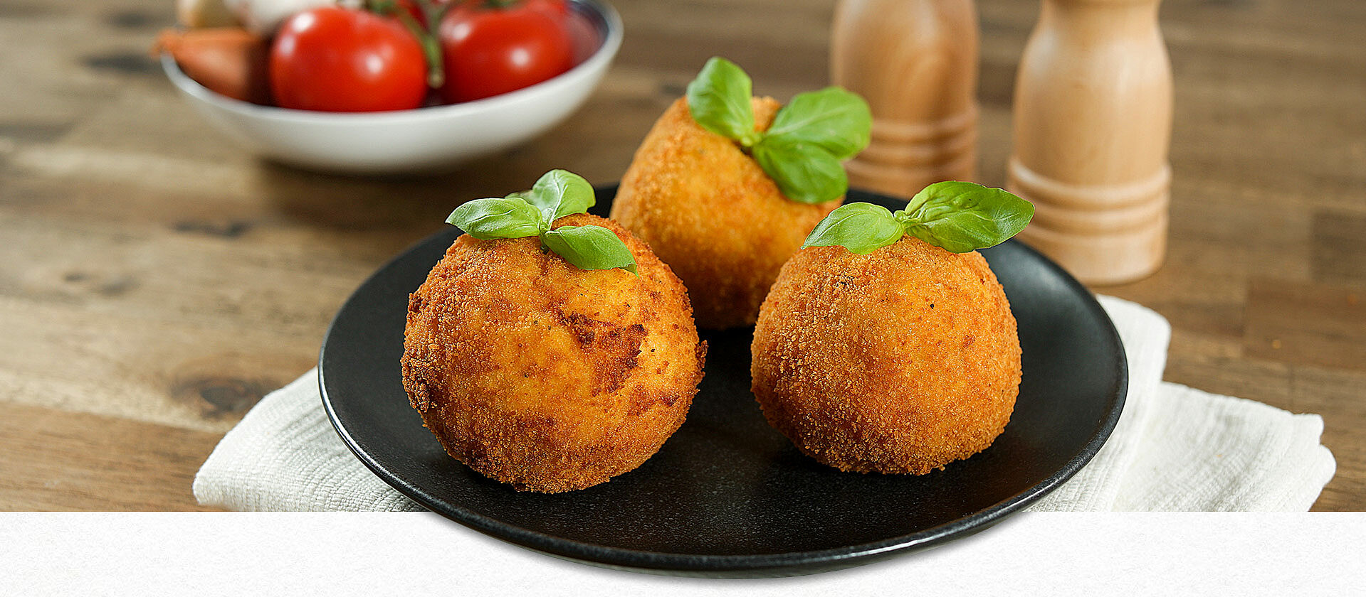 Arancini di riso - Reisbällchen auf einem schwarzen Teller. Im Hintergrund Salz- und Pfefferstreuer sowie frische Tomaten.