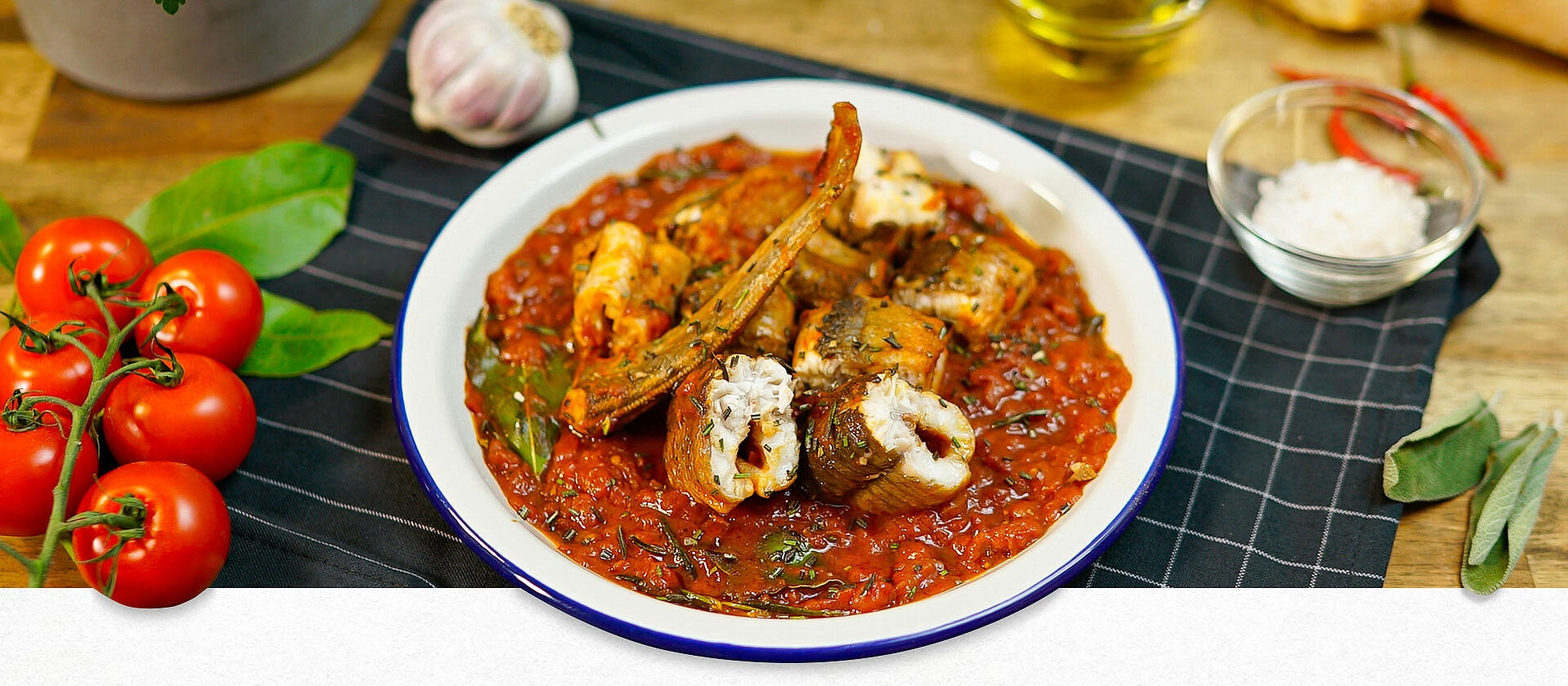 Auf einem weißen Teller mit blauen Rand ist ein traditionelles italienisches Aal-Rezept zu sehen. Der Aal liegt in Tomatensoße. Verschiedene frische Zutaten wie Tomaten, Basilikum sind als Deko zu sehen.