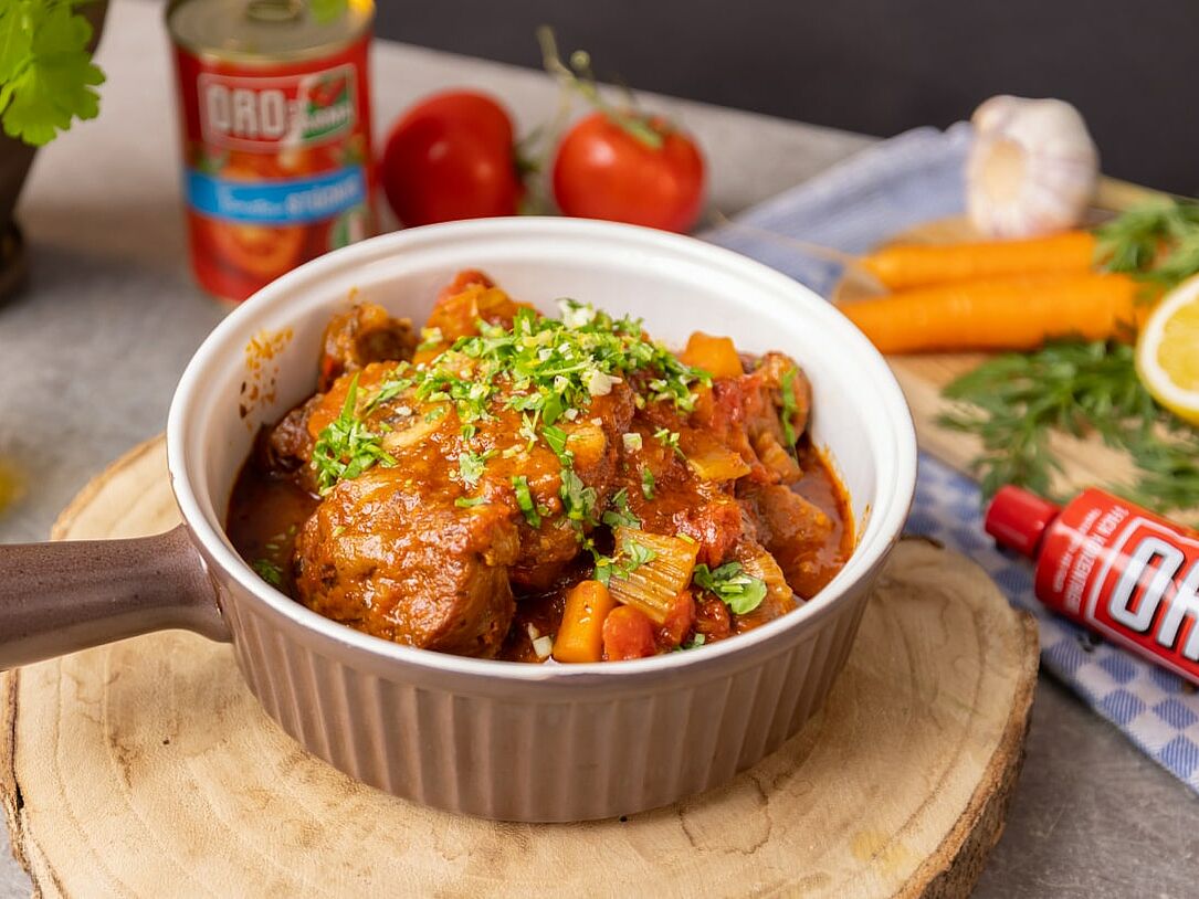 In einem Schmortopf ist Ossobuco alla milanese anrichtet. Der Topf steht auf einem runden Holzbrett. Im Hintergrund verschiedene frische Zutaten. 