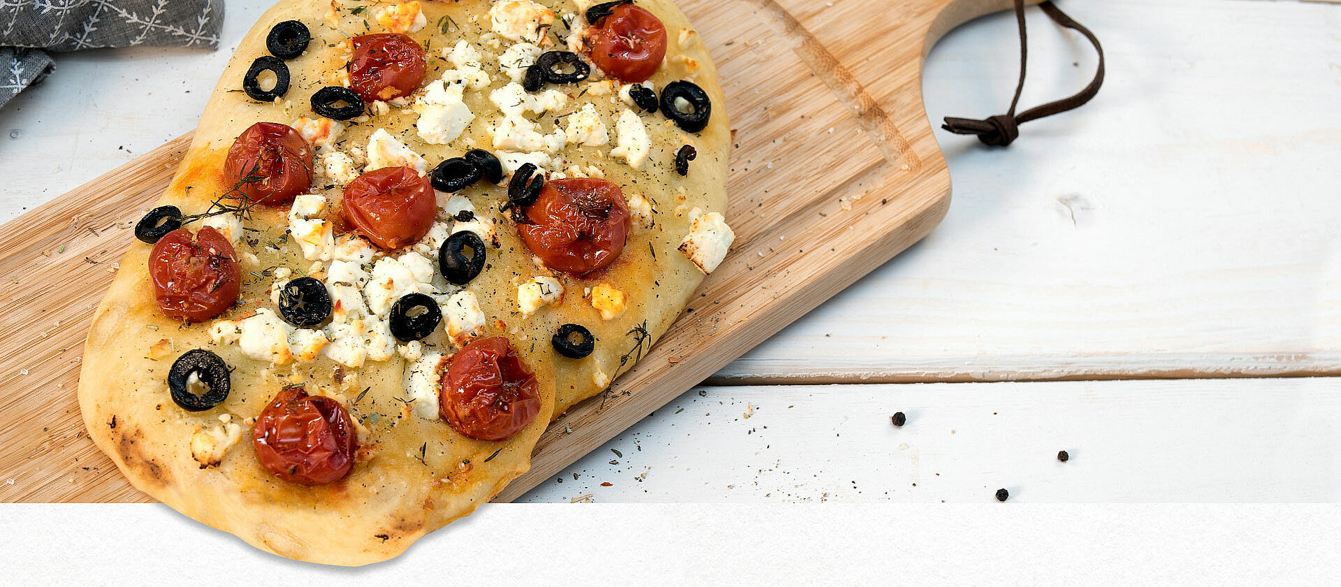 Auf einem Holzbrett liegt eine Focaccia mit Tomaten, Oliven und Schafskäse