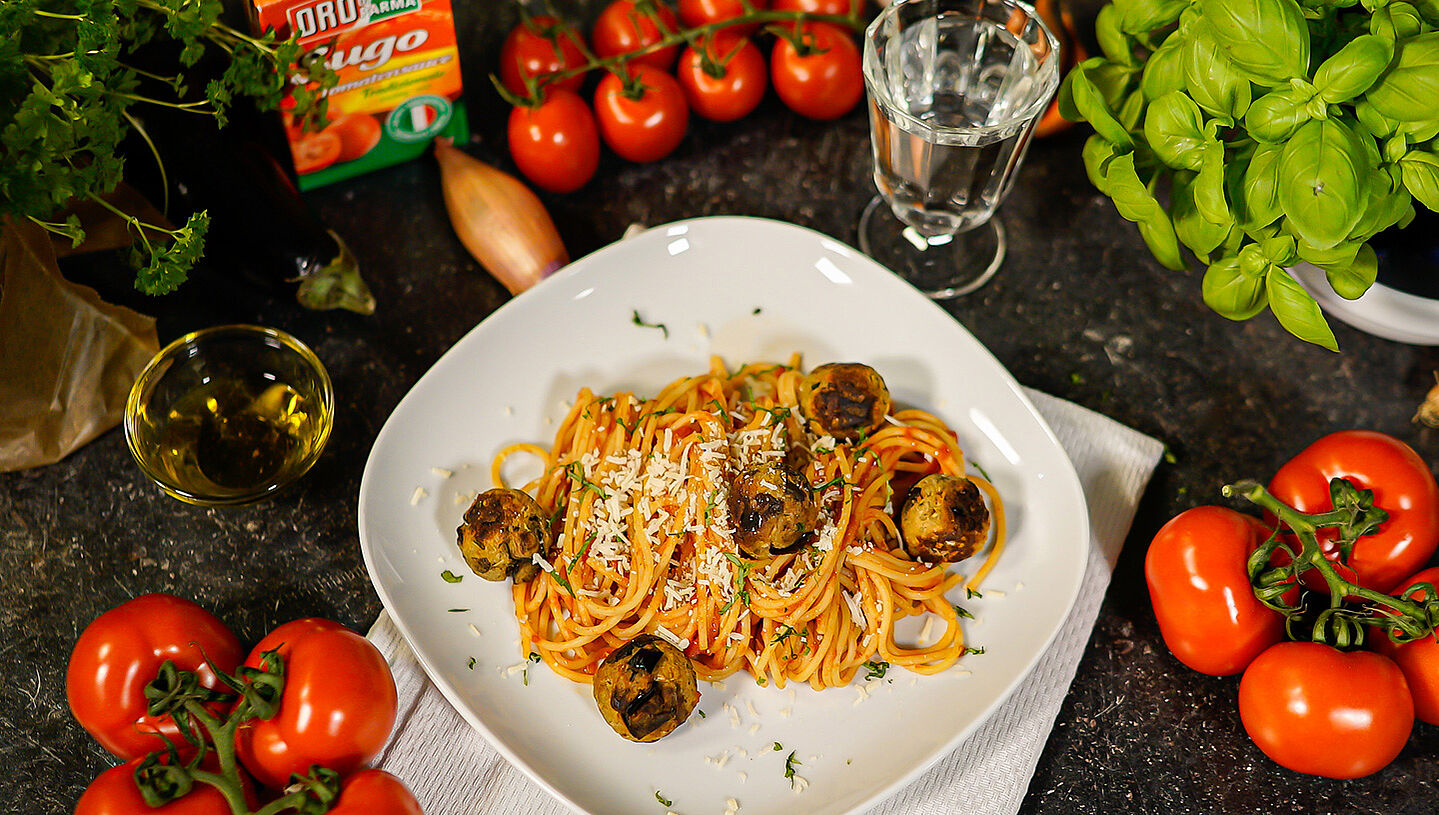 Auf einem weißen Teller sind Spaghetti mit Auberginenbällchen angerichtet. Daneben liegen frische Tomaten und Basilikum.