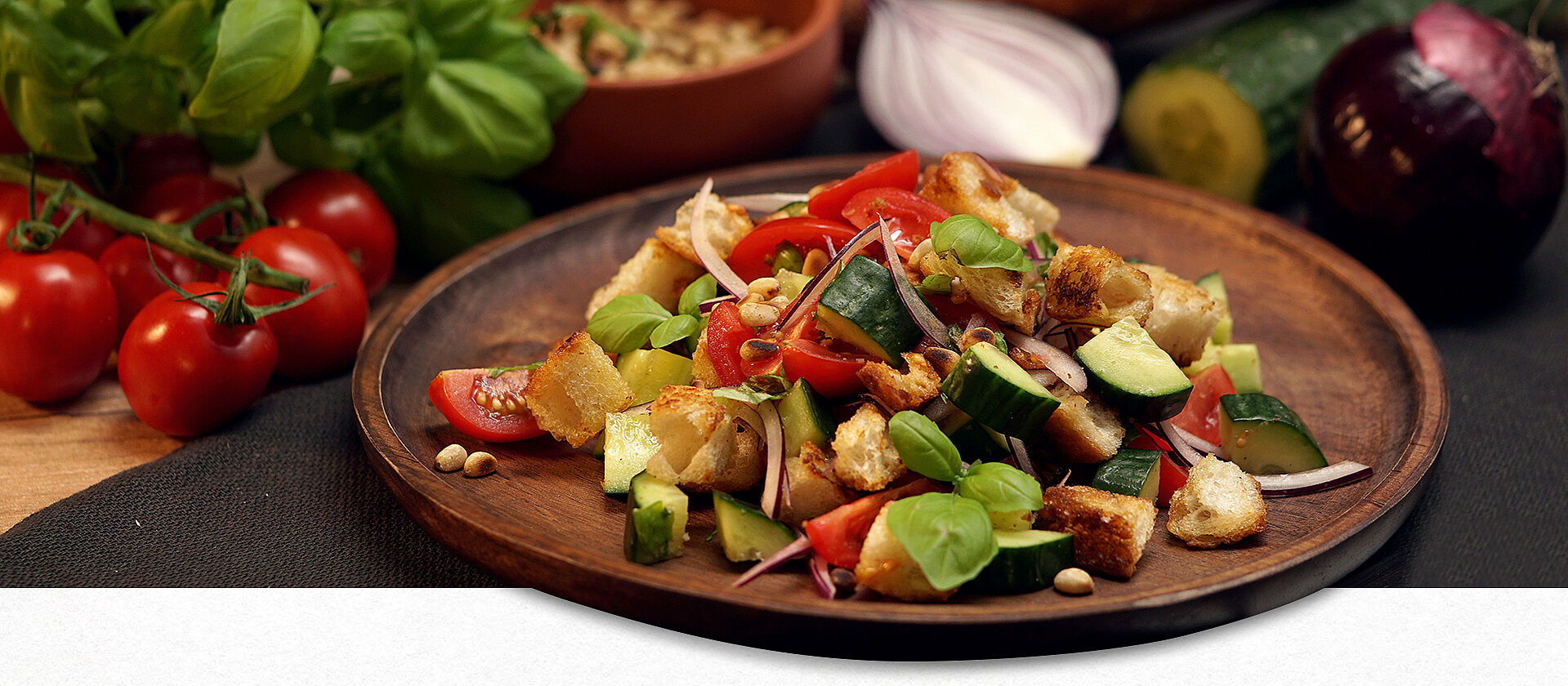 Auf einem Holzteller ist der italienischer Brotsalat namens Panzanella mit Gurken, Tomaten, Zwiebeln angerichtet