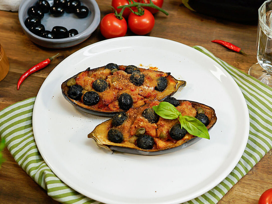 Auf einem weißen Teller liegen zwei Auberginenhälften. Beide sind gefüllt und überbacken. Im Hintergrund stehen ORO di Parma Tomaten, Oliven und frische Tomaten.