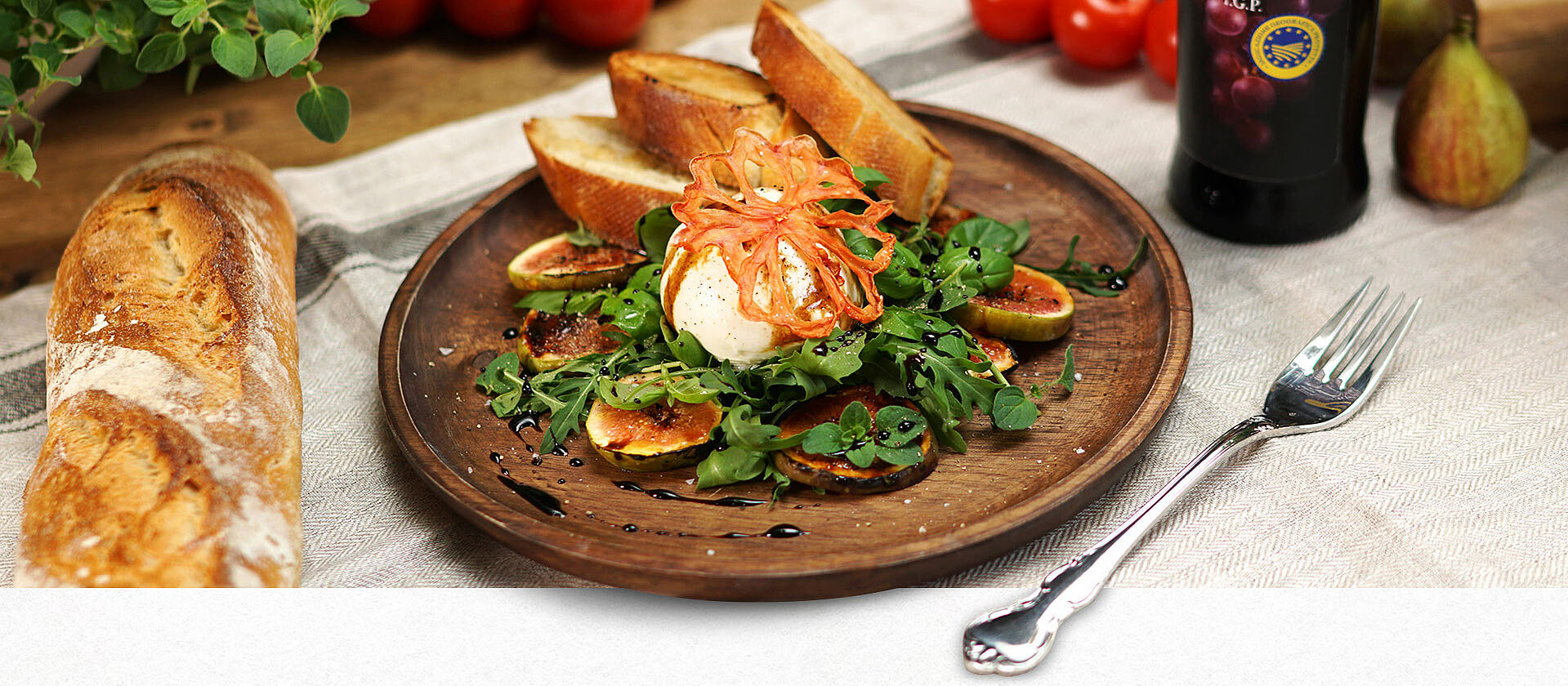 Auf einem Holzteller ist ein Salat mit Burrata und Feigen sowie Ciabatta Brot angerichtet.