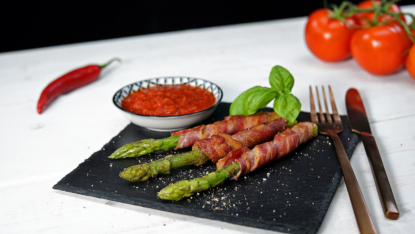 Grüner Spargel im Speckmantel angerichtet auf einer Schieferplatte mit Tomaten Sugo