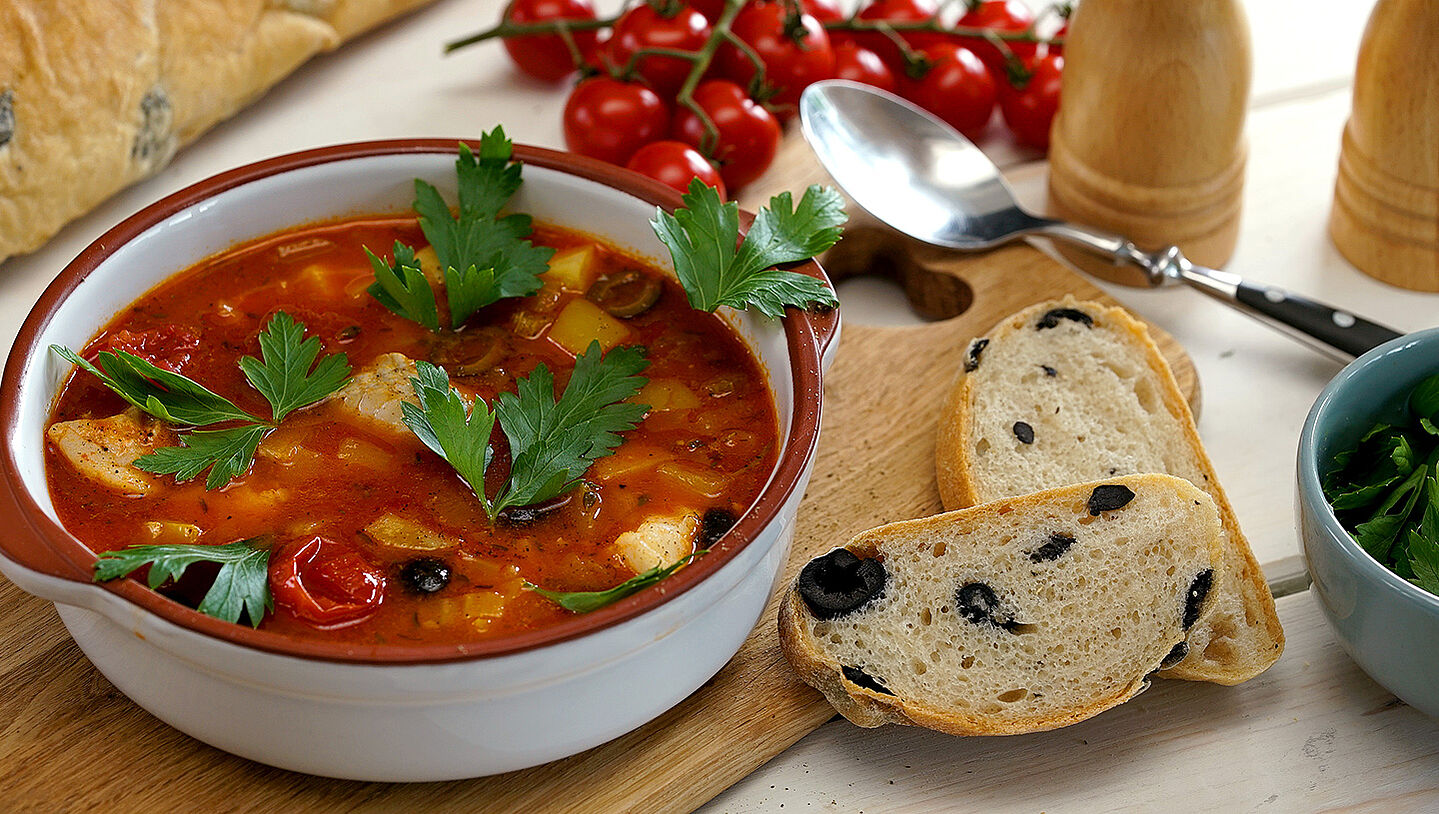 Eine Schüssel mit Tomaten-Fischsuppe, daneben zwei Scheiben Olivenbrot