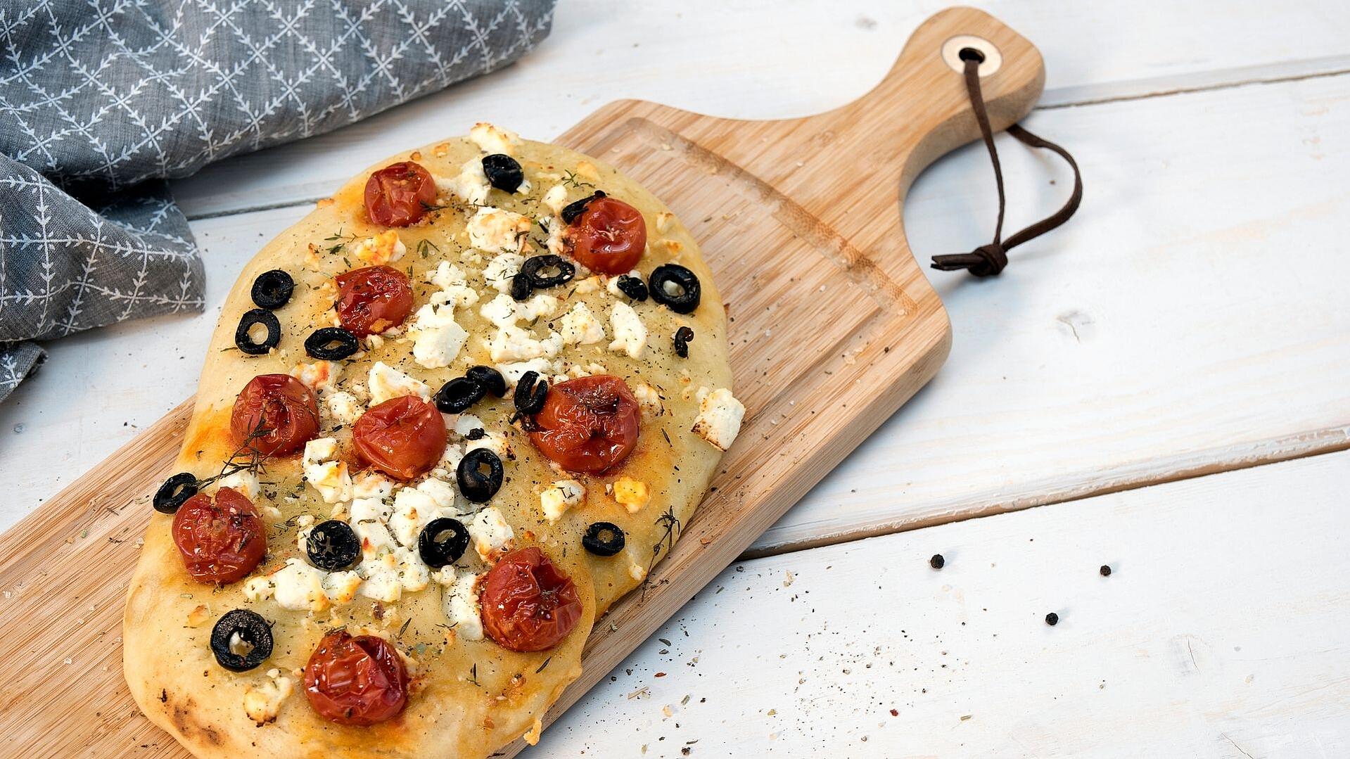 Auf einem Holzbrett liegt eine Focaccia mit Tomaten, Oliven und Schafskäse