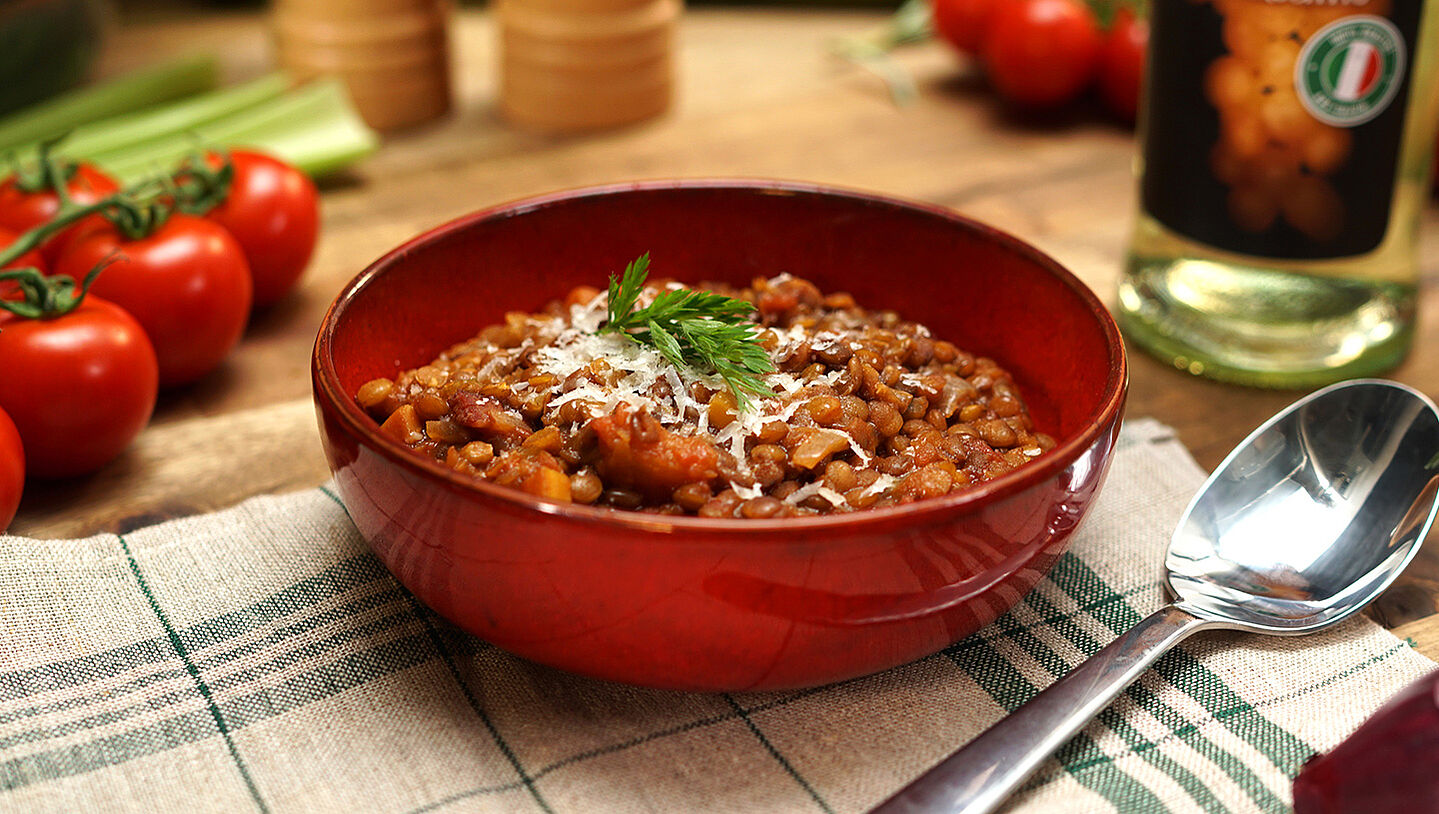 In einer Suppenschüssel ist ein italienischer Linseneintopf. Im Hintergrund liegen Tomaten, ein Salz- und Pfefferstreuer und eine Flasche Essig.