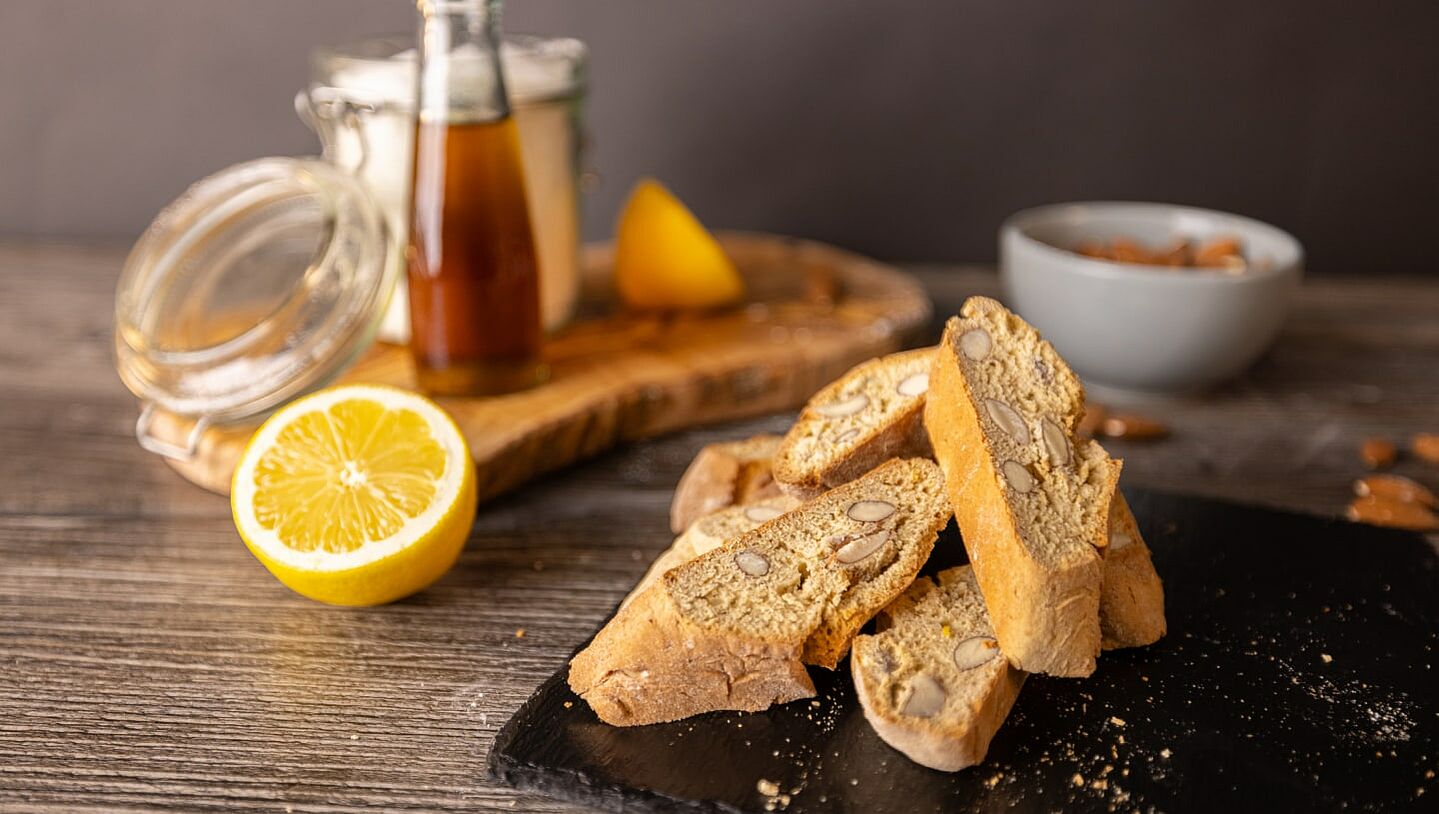 Auf einer Schieferplatte liegen 4 italienische Cantuccini. Im Hintergrund eine Tasse Kaffee.