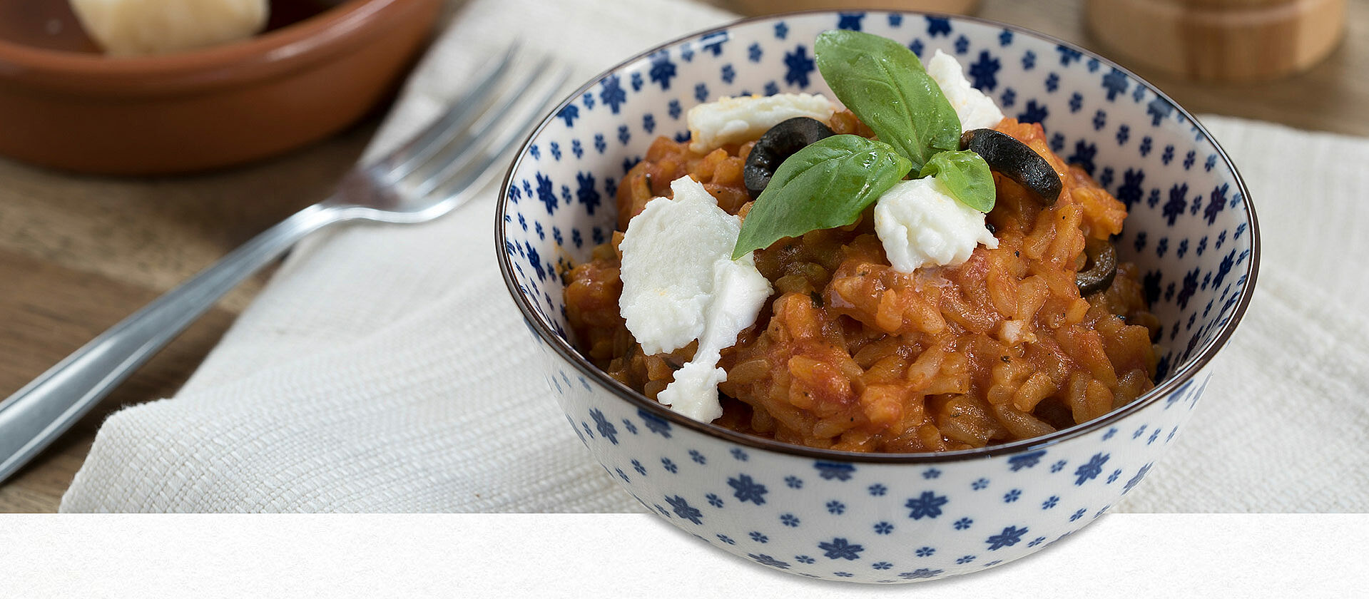 Alternativ-Text: Eine Schale gefüllt mit Tomaten-Risotto, garniert mit Oliven, Mozzarella und Basilikum