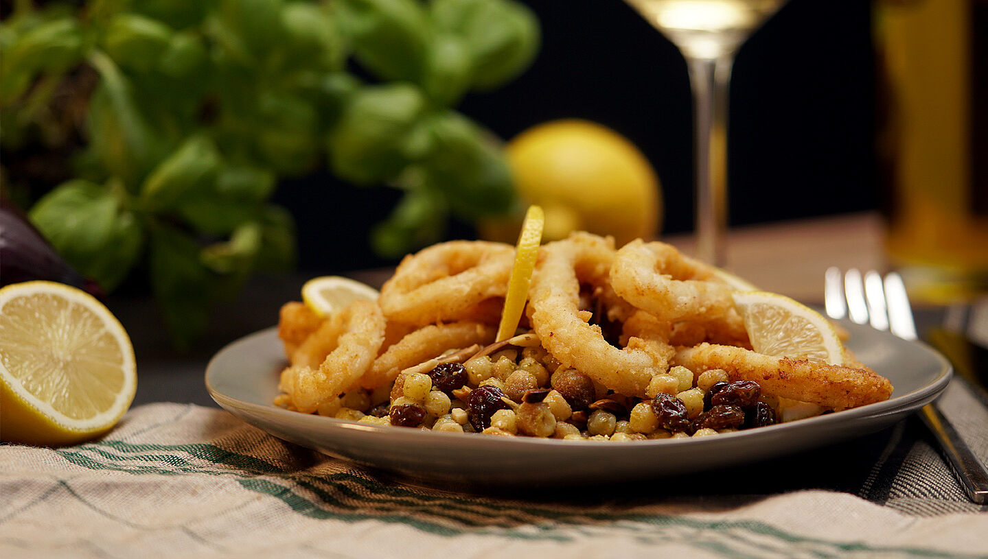 Ein Teller mit Fregola sarda tostata und frittierten Calamari. Im Hintergrund eine halbe Zitrone und frischer Basilikum.
