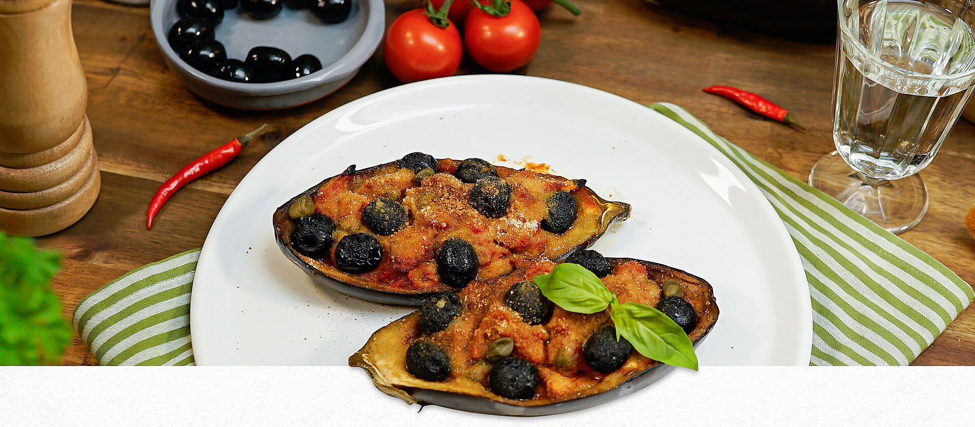 Auf einem weißen Teller liegen zwei Auberginenhälften. Beide sind gefüllt und überbacken. Im Hintergrund stehen ORO di Parma Tomaten, Oliven und frische Tomaten.