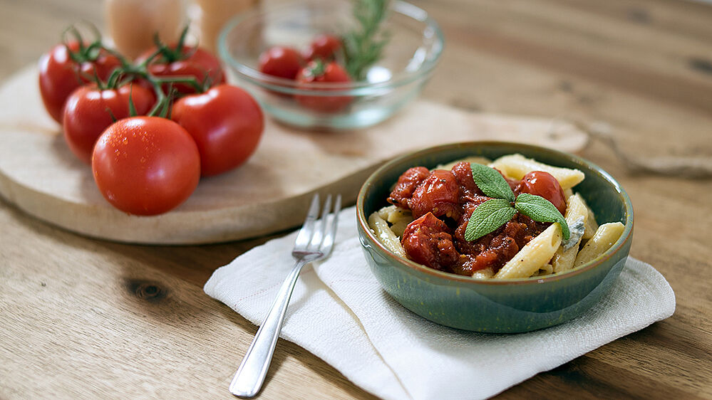 Eine Schüssel mit Penne Gorgonzola, links daneben ein Holzbrett mit Tomaten