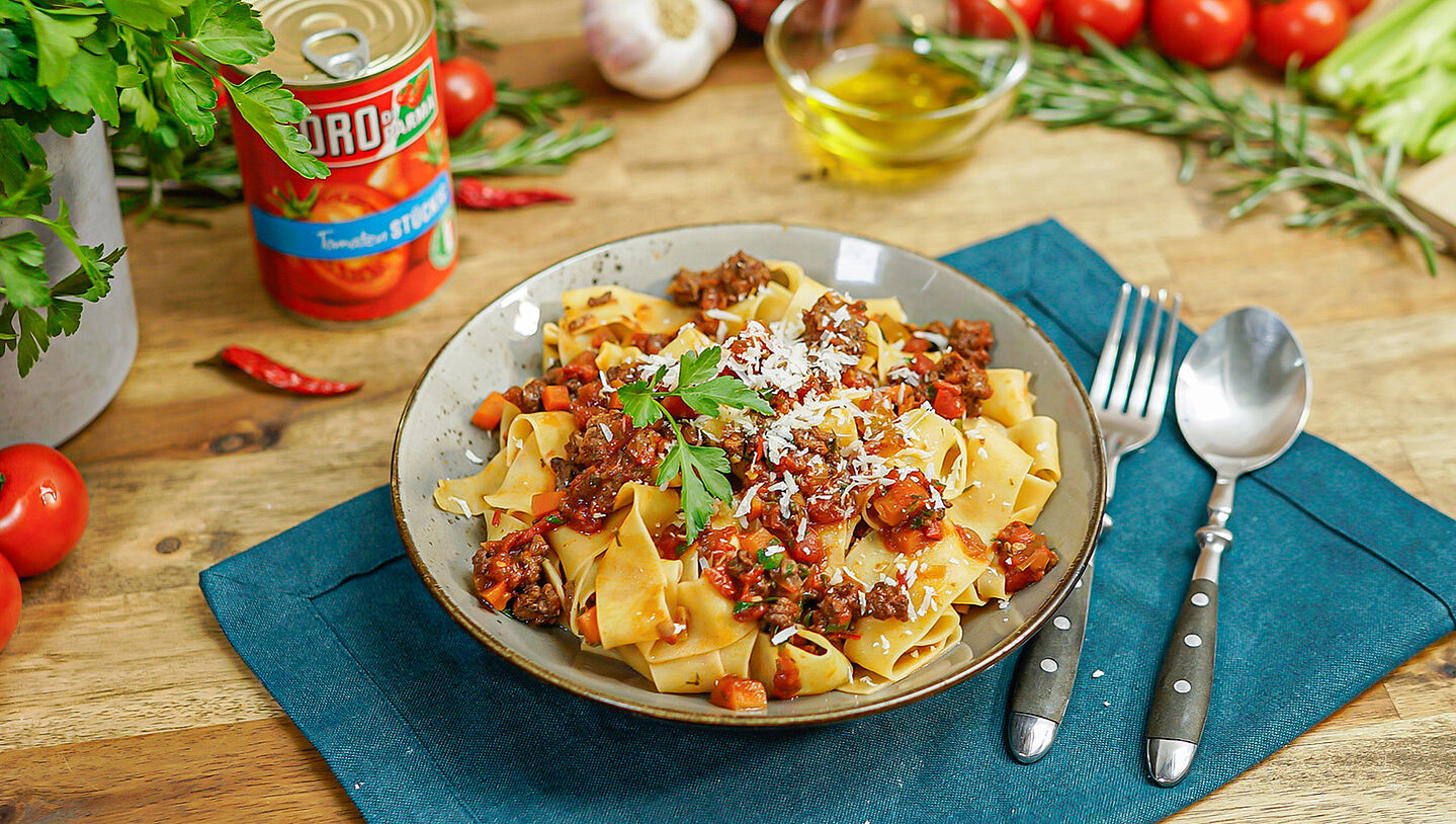 Ein Teller gefüllt mit Pappardelle und Wildschwein-Ragú steht auf einem blauen Tischset. Im Hintergrund Basilikum und eine Dose ORO di Parma stückige Tomaten.