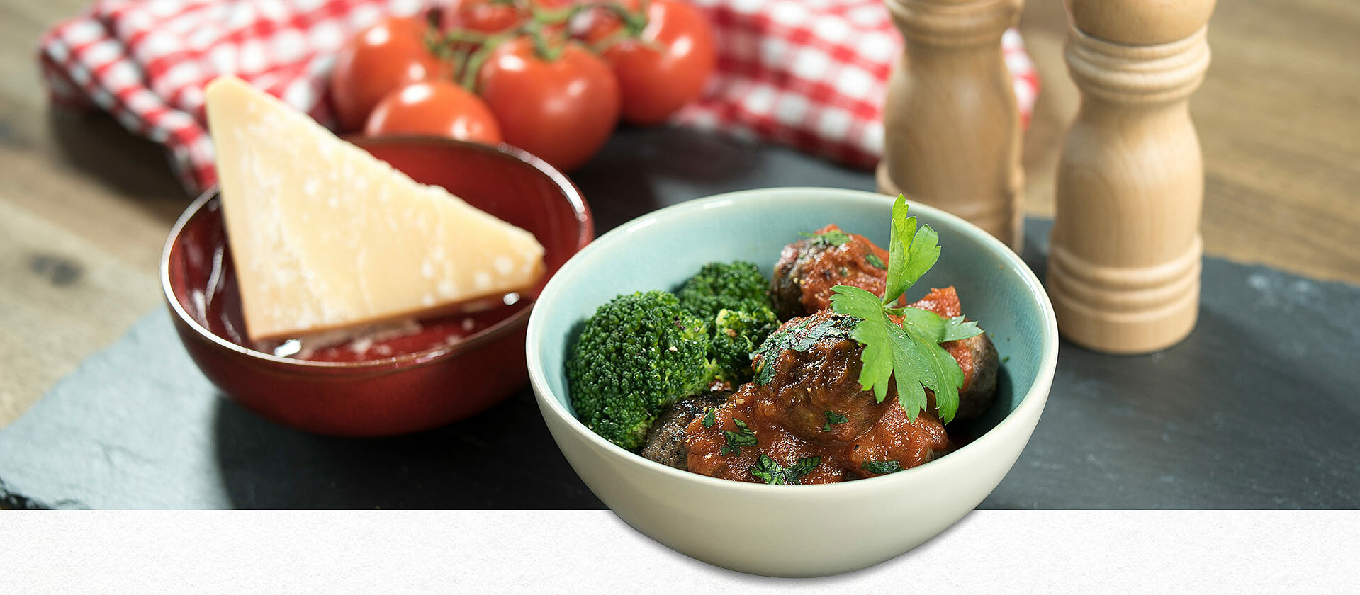Eine Schüssel mit Salami-Fleischbällchen in Tomatensugo mit Brokkoli, daneben eine Schüssel mit einem Stück Parmesan