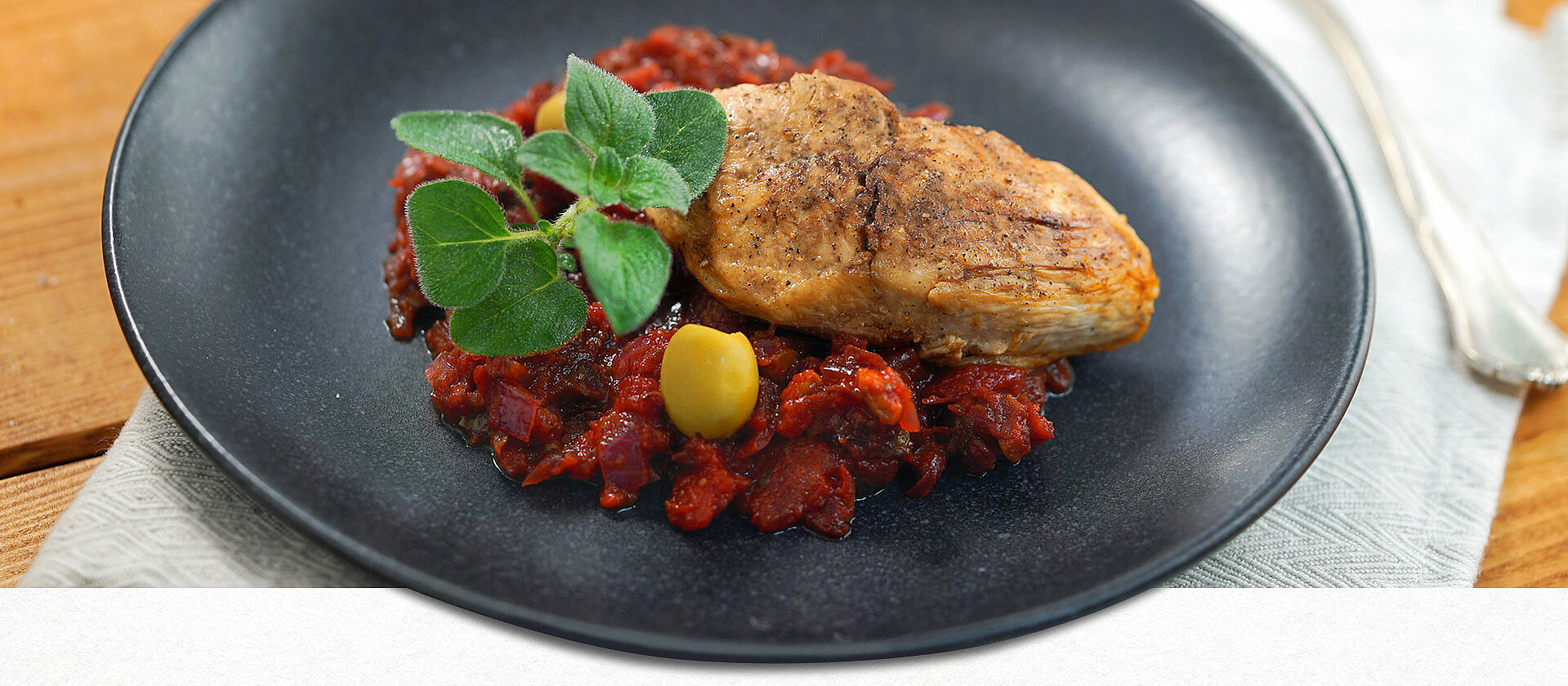 Ein schwarzer Teller mit Hähnchenbrust auf Tomaten-Oliven-Sauce 