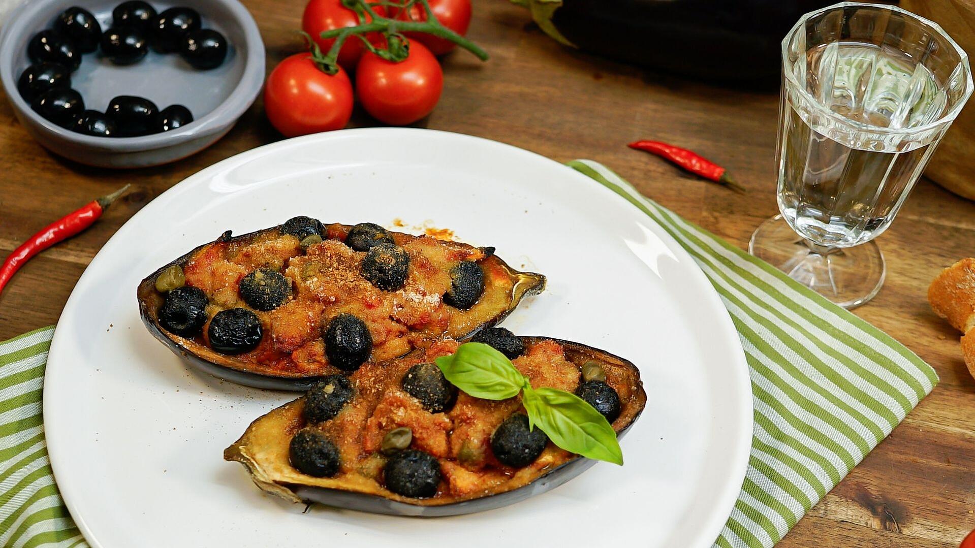 Auf einem weißen Teller liegen zwei Auberginenhälften. Beide sind gefüllt und überbacken. Im Hintergrund stehen ORO di Parma Tomaten, Oliven und frische Tomaten.
