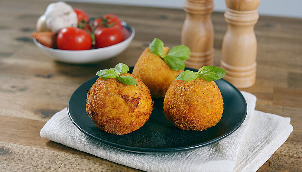 Arancini di riso - Reisbällchen auf einem schwarzen Teller. Im Hintergrund Salz- und Pfefferstreuer sowie frische Tomaten.