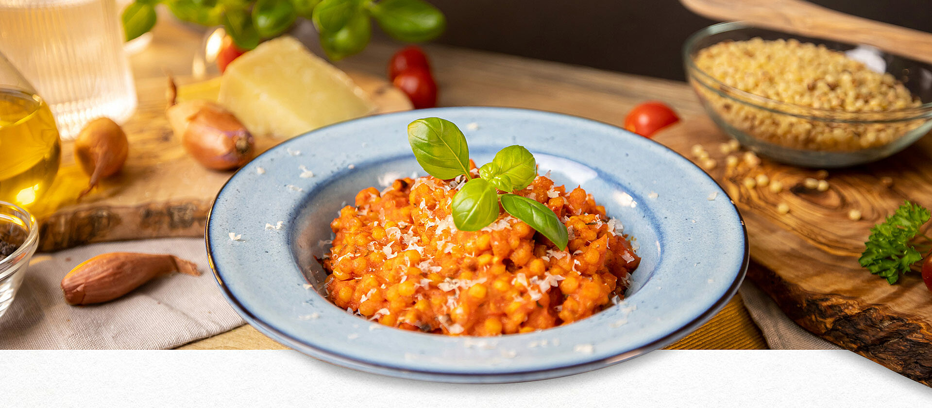 In einem hellblauen Teller sind Fregola Sarda mit Tomatensauce angerichtet. Im Hintergrund sind Parmesan, Zwiebeln, frischer Basilikum erkennbar. 