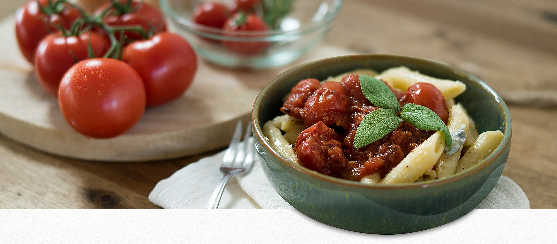 Eine Schüssel mit Penne Gorgonzola, links daneben ein Holzbrett mit Tomaten