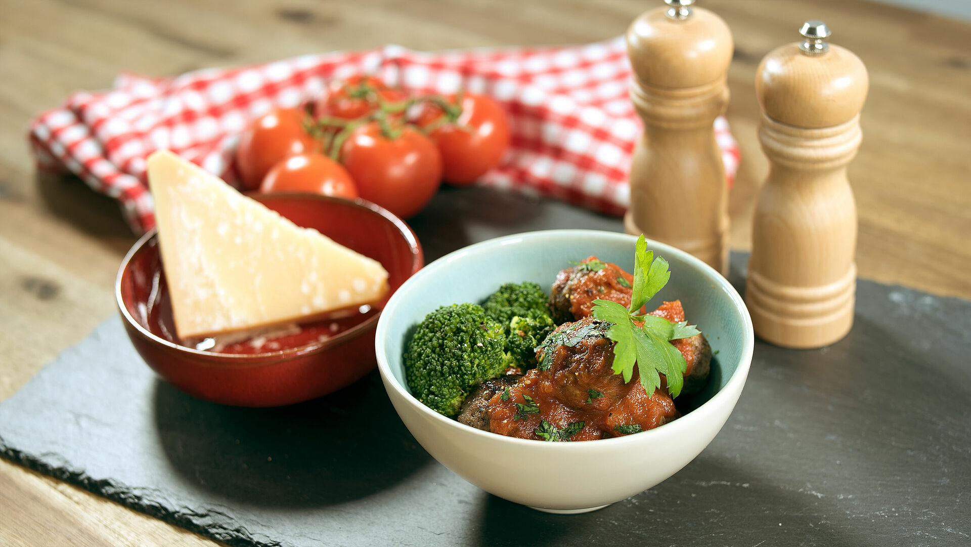 Eine Schüssel mit Salami-Fleischbällchen in Tomatensugo mit Brokkoli, daneben eine Schüssel mit einem Stück Parmesan