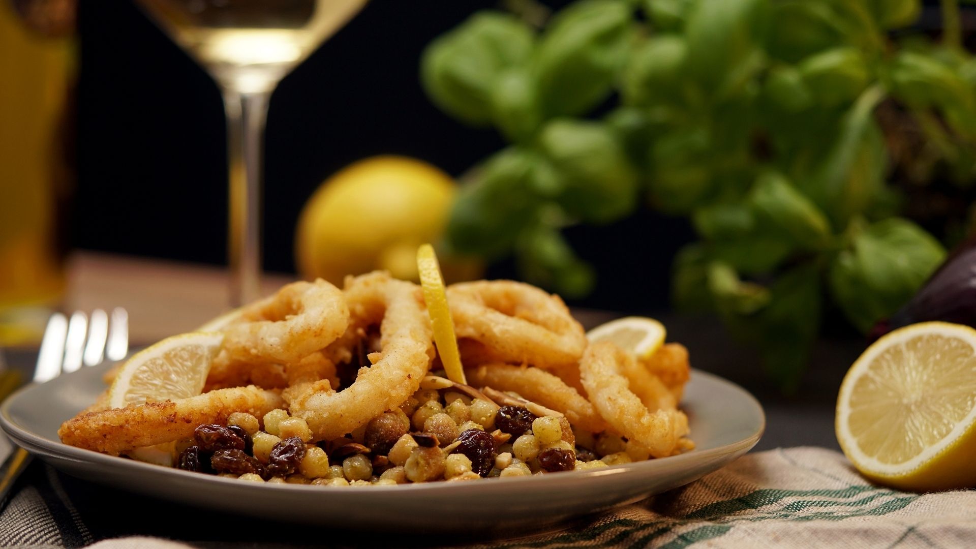 Ein Teller mit Fregola sarda tostata und frittierten Calamari. Im Hintergrund eine halbe Zitrone und frischer Basilikum.