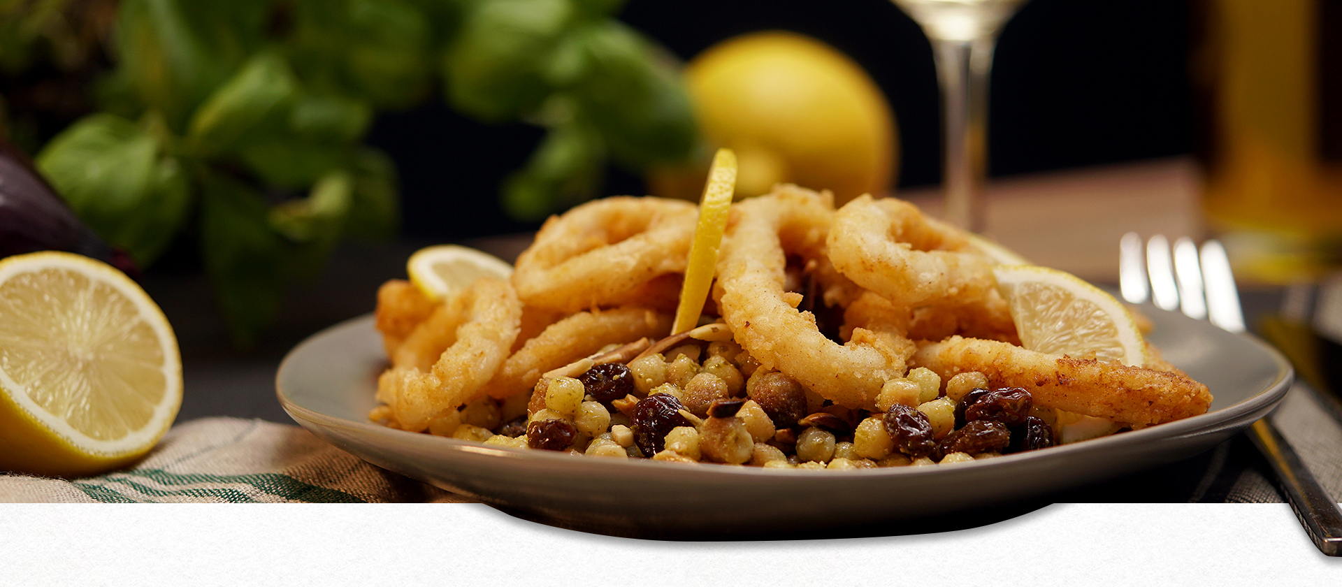 Ein Teller mit Fregola sarda tostata und frittierten Calamari. Im Hintergrund eine halbe Zitrone und frischer Basilikum.