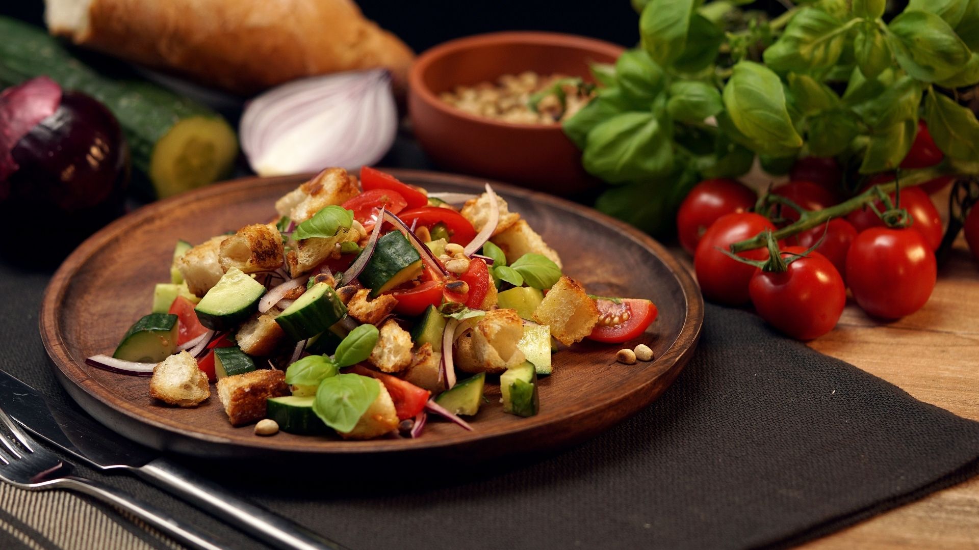 Auf einem Holzteller ist der italienischer Brotsalat namens Panzanella mit Gurken, Tomaten, Zwiebeln angerichtet