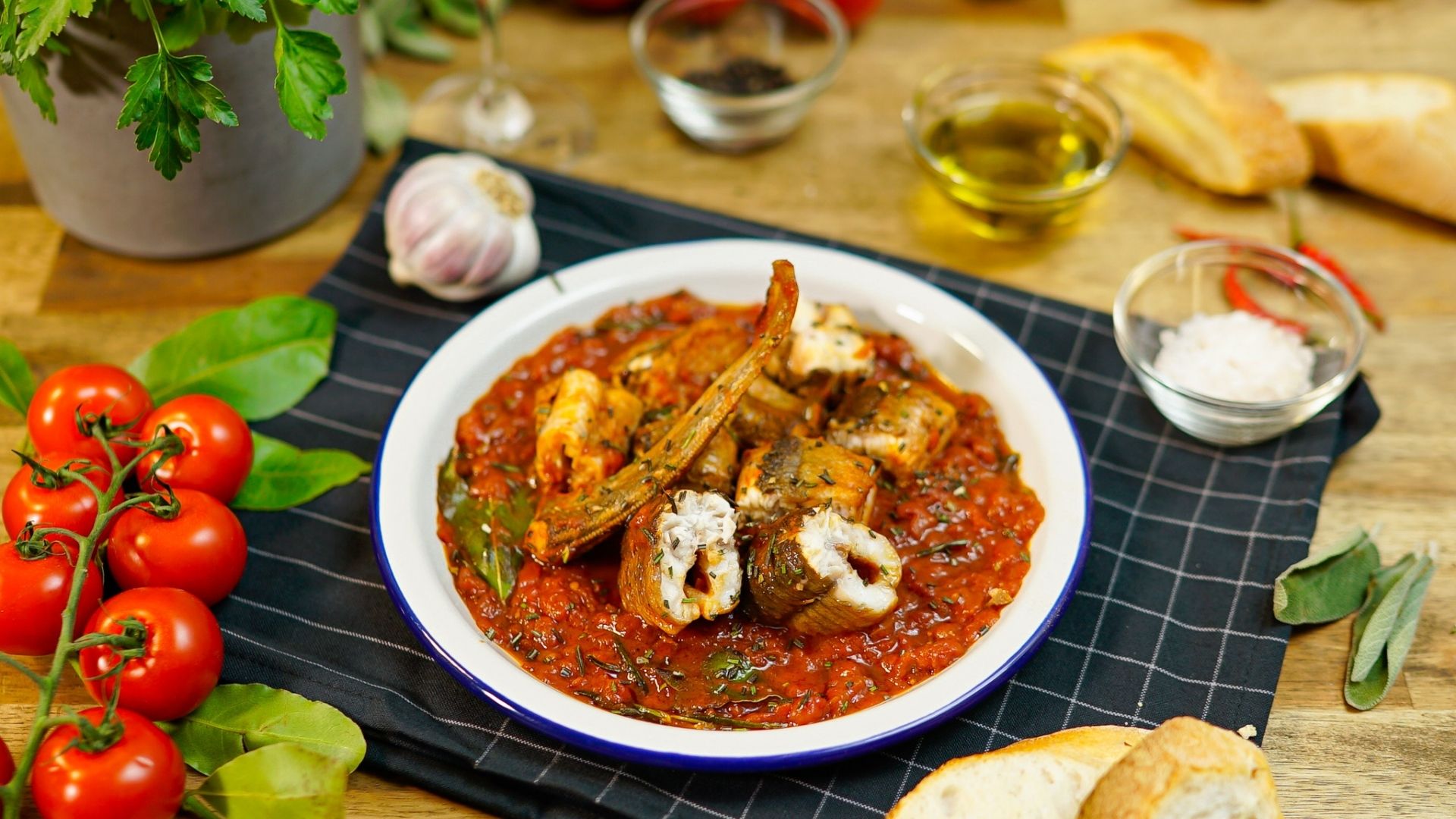 Auf einem weißen Teller mit blauen Rand ist ein traditionelles italienisches Aal-Rezept zu sehen. Der Aal liegt in Tomatensoße. Verschiedene frische Zutaten wie Tomaten, Basilikum sind als Deko zu sehen.