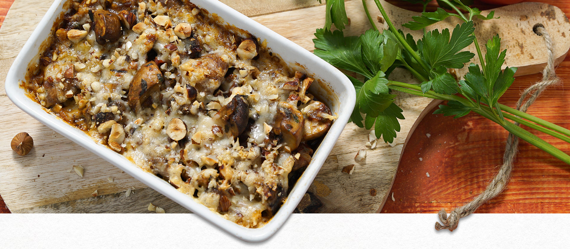 Eine kleine, weiße Auflaufform mit gratiniertem Gemüse. Daneben liegt Petersilie und eine Tomate.