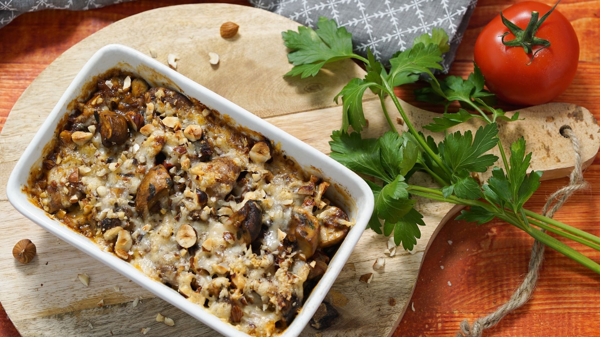 Eine kleine, weiße Auflaufform mit gratiniertem Gemüse. Daneben liegt Petersilie und eine Tomate.