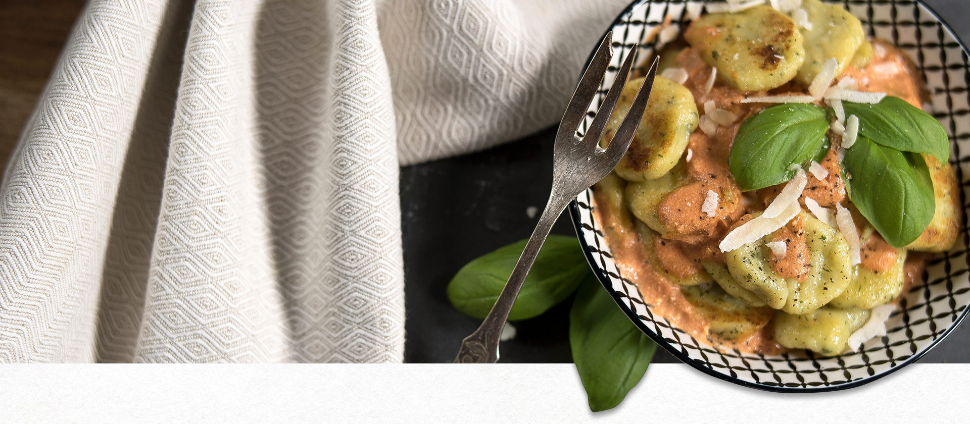 Basilikum-Gnocchi angerichtet auf einem weißen Teller mit schwarzem Karo-Muster, daneben eine graue Serviette