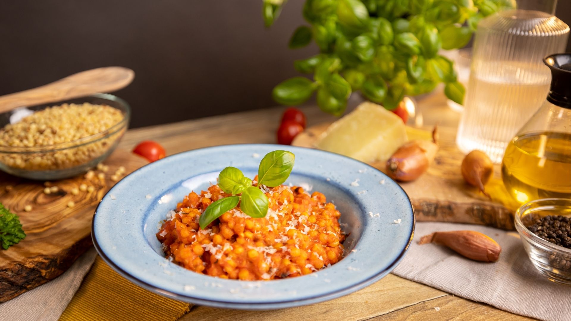 In einem hellblauen Teller sind Fregola Sarda mit Tomatensauce angerichtet. Im Hintergrund sind Parmesan, Zwiebeln, frischer Basilikum erkennbar. 
