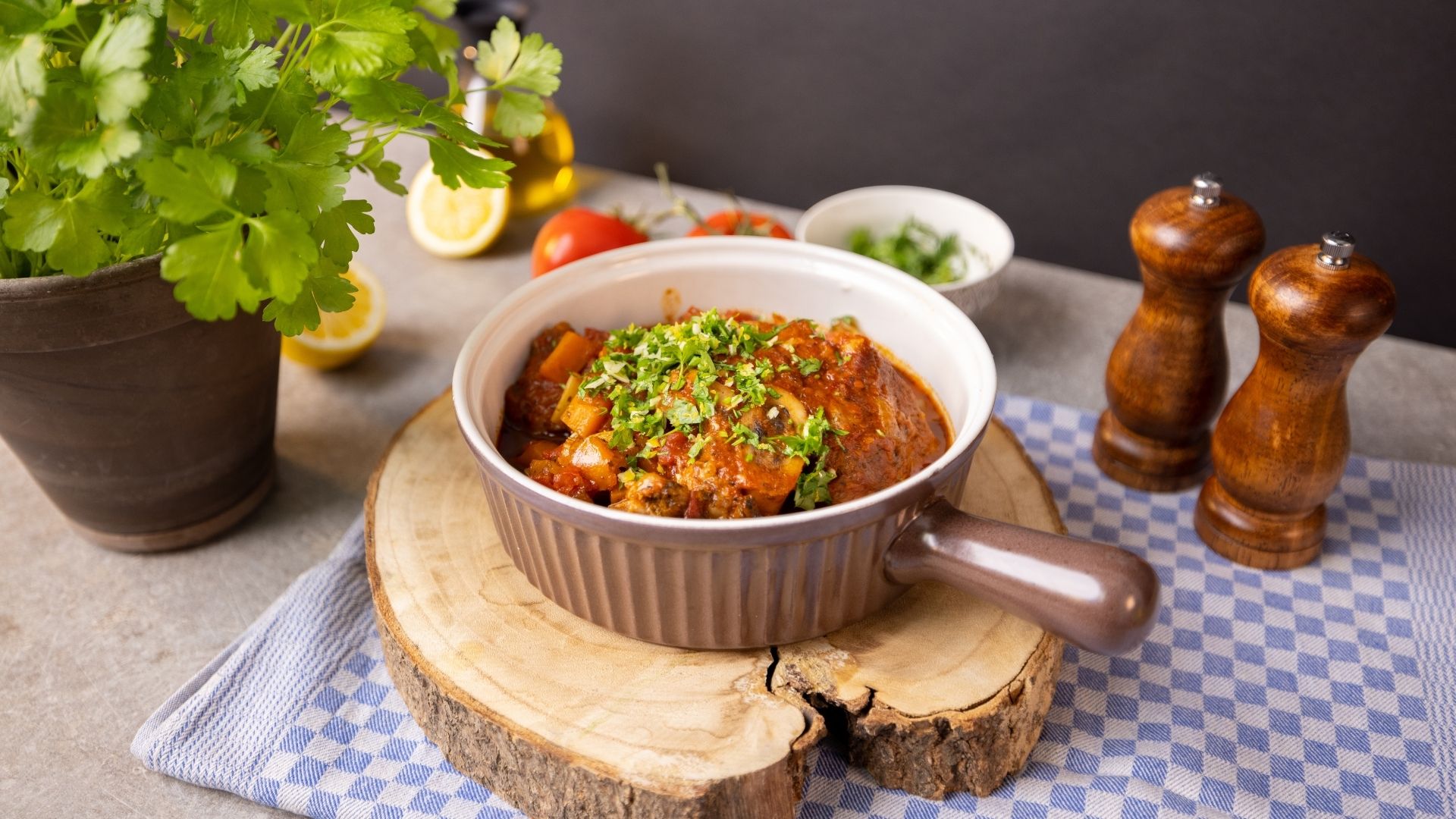 In einem Schmortopf ist Ossobuco alla milanese anrichtet. Der Topf steht auf einem runden Holzbrett. Im Hintergrund verschiedene frische Zutaten. 